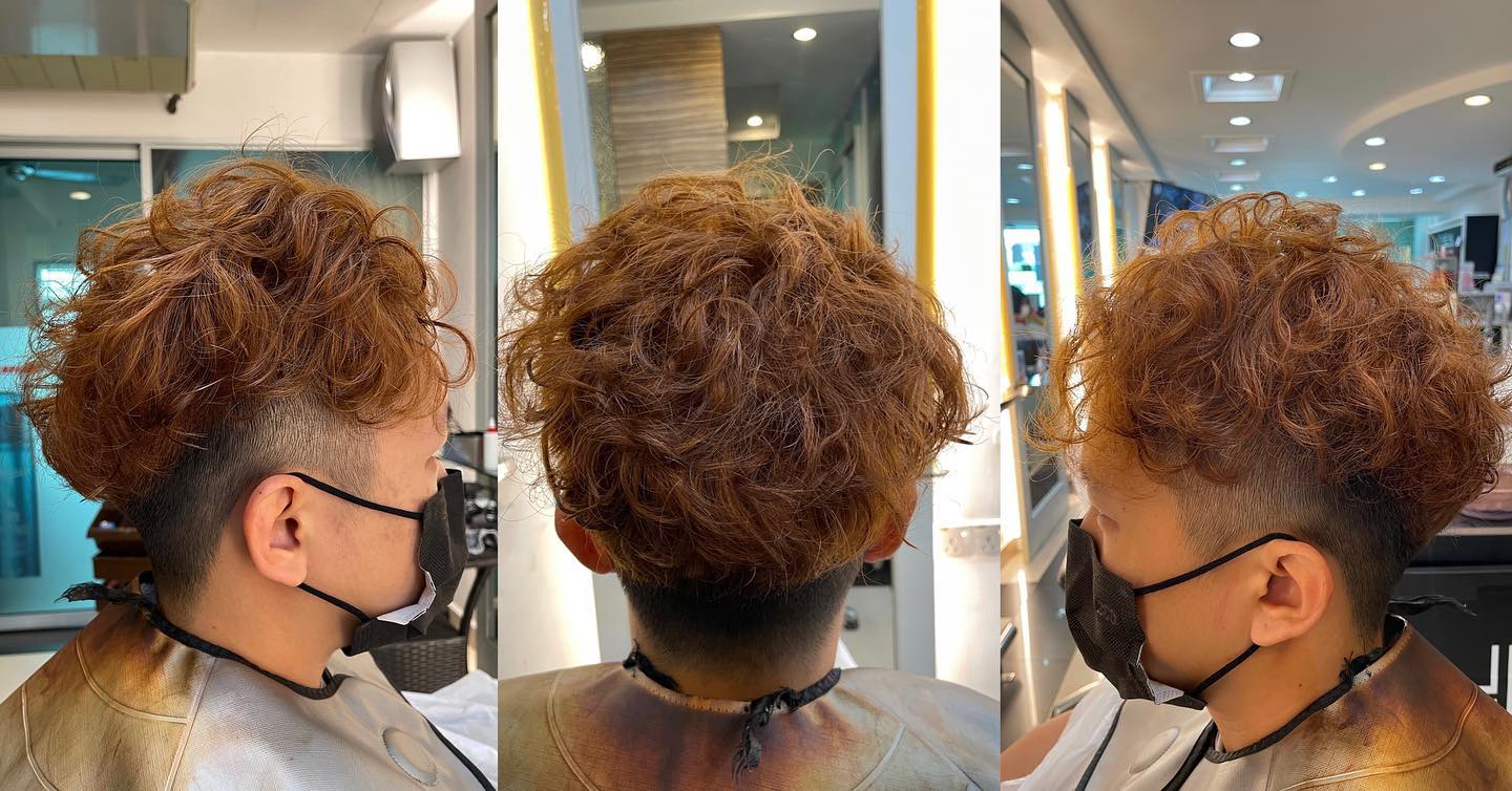 Person with curly light brown hair styled in a voluminous modern cut, sitting in a salon chair wearing a protective cape and black face mask, surrounded by bright salon lighting and mirrors, creating a professional and welcoming atmosphere